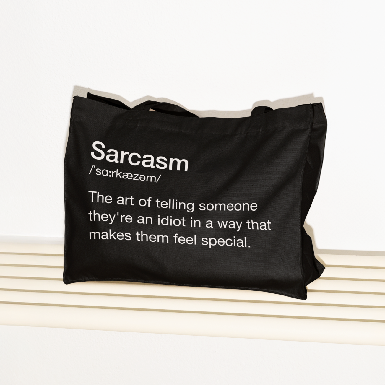 Juodas tote bag'as su baltu užrašu „Sarcasm – The art of telling someone they're an idiot...“, padėtas ant šviesaus suolo.